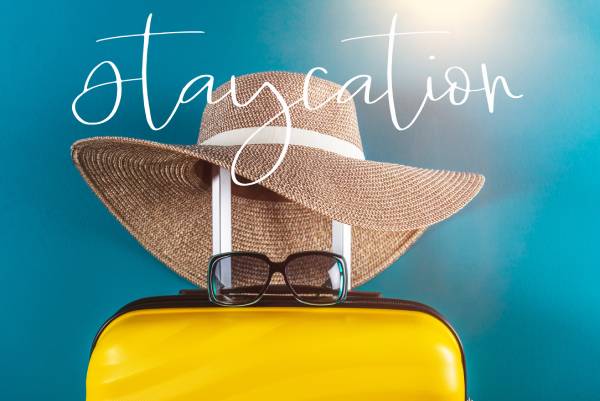 Staycation word. Bright and stylish cabin size suitcase with straw hat against bright blue background. Easy travel at home country during coronavirus outbreak with little baggage concept.